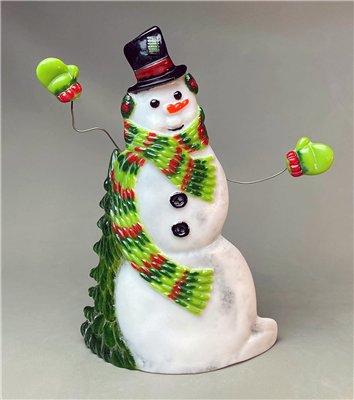 A fused glass snowman displayed on grey background. The snowman has a black top hat with a red ribbon, and green and red earmuffs and mittens. He also has a long scarf with stripes of red, dark green, and lime green. He has two black buttons on his chest.