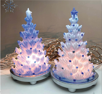 Two fused glass Christmas trees displayed on silver bases with lights behind them. The left tree is frosty blue with dark blue ornaments and a white star on top, and the right tree is white with various blue ornaments and a blue star.