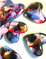 Glass hearts on a cream background. The hearts are made with a variety of glass, and have patches of dark brown, clear, red, yellow, and opalescent blue. A hand holds one heart closer to the camera in the top left of the image.