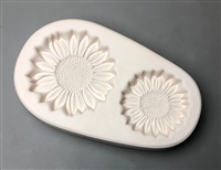 An ovoid white ceramic mold for fusing hot glass on a grey background. Two separate detailed sunflowers have been carved into it. The left flower is slightly larger than the right.