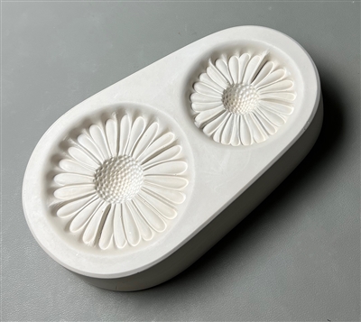 An ovoid white ceramic mold for fusing hot glass on a grey background. Two separate detailed coneflowers with deep centers have been carved into it. The left flower is slightly larger than the right.