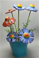 A bouquet of fused glass flowers arranged on green wooden stems in a blue bucket filled with greenery. The flowers are purple or red with yellow centers. Some flowers have been left flat while others have been shaped into more natural shapes.