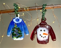 Two fused glass sweater ornaments hang from a branch with small lights wrapped around it. The left sweater is blue with white trim and a colorful Christmas tree design. The right sweater is red with painted gold trim and a large smiling snowman face.