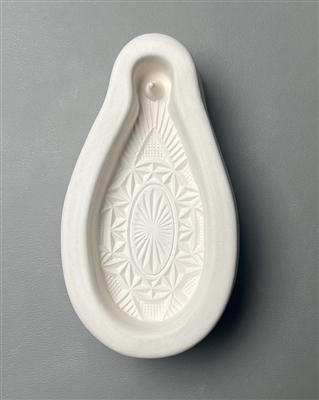 A white ceramic mold for fusing hot glass on a grey background. A detailed teardrop ornament with a faceted crystal pattern has been carved into it. The top of the ornament has a post to allow for hanging after fusing.
