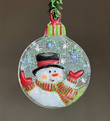 A round, fused glass ornament hanging in front of a grey background. The ornament has a smiling snowman on an iridescent clear background with silver and white snowflakes. He has a green and red striped scarf as does the top of the ornament.