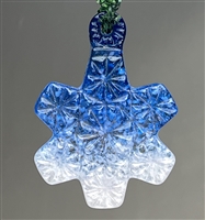 A fused glass snowflake ornament hanging in front of a grey background. The ornament has an intricate cut crystal design all over it and is filled with a color gradient going from dark blue at the top to white at the bottom.