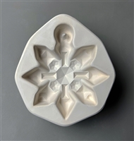 A white ceramic mold for fusing hot glass on a grey background. An eight-armed snowflake ornament with a faceted and geometric design has been carved into it. The top of the snowflake has a post to allow for hanging after fusing.