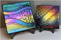 Two fused glass easels hold up two different glass squares filled with designs. The larger left easel has a square with lines and crosses, while the smaller right square has hatches and spirals. The glass is all dark and iridescent, reflecting many colors