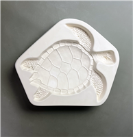 A white ceramic mold for fusing hot glass on a grey background. A large and detailed top-down view of a sea turtle facing right has been carved into it.