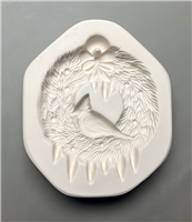 A white ceramic mold for fusing glass displayed on a grey background. A wreath made of evergreen branches has been carved into it. A cardinal is carved sitting in the middle of the wreath, and there are a few icicles at the bottom and a bow at the top.