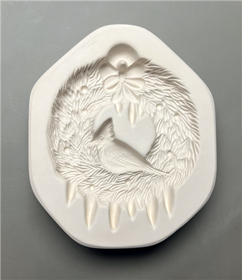 A white ceramic mold for fusing glass displayed on a grey background. A wreath made of evergreen branches has been carved into it. A cardinal is carved sitting in the middle of the wreath, and there are a few icicles at the bottom and a bow at the top.