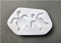 A white ceramic mold for fusing hot glass on a grey background. The top-down view of two small sea turtles has been carved into it. Their fins are outstretched and heads curved slightly, as if swimming.