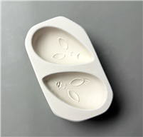 A white ceramic mold for fusing glass on a grey background. Two teardrop-shaped alien faces have been carved into it, oriented in opposite directions. Both faces have large, slanted oblong eyes and small mouths, and the bottom face has lips and eyelashes