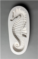 A white ceramic mold for fusing glass in the shape of an elongated oval. A large detailed seahorse is carved into it. The seahorse faces left, with long spines atop its head that get shorter down its back. It has a curled tail and prominent dorsal fin.