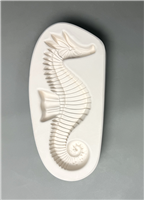 A white ceramic mold for fusing glass in the shape of an elongated oval. A detailed seahorse is carved into it. The seahorse faces right, with long spines atop its head that get shorter down its back. It has a curled tail and prominent dorsal fin.