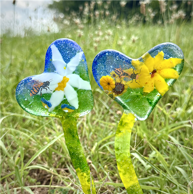 Two stakes made of fused glass displayed in a grassy field. The stakes each have thin curved stems and are topped with slightly off center hearts. They are made with shades of green and blue glass, and each has several flower shapes in its heart portion.