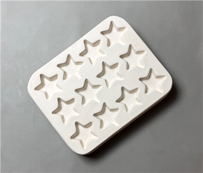 A rectangular white ceramic mold for fusing glass on a grey background. Twelve identical, evenly spaced stars have been carved into it. Each star has five points, and the tips of each point are very slightly rounded.