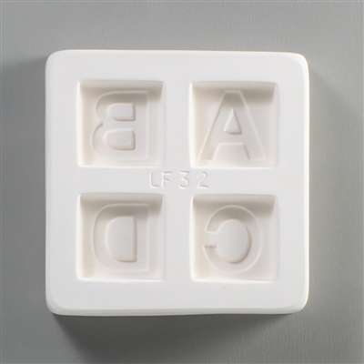 A square white ceramic mold for fusing hot glass on a grey background. Four separate but identically sized squares have been carved into it with equal space between them. Each square has one of the letters A, B, C, or D carved in block print in it.