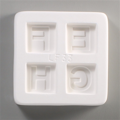 A square white ceramic mold for fusing hot glass on a grey background. Four separate but identically sized squares have been carved into it with equal space between them. Each square has one of the letters E, F, G, or H carved in block print in it.