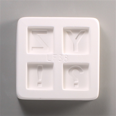 A square white ceramic mold for fusing hot glass on a grey background. Four separate but identically sized squares have been carved into it with equal space between them. Each square has one of the characters Y, Z, !, or ? carved in block print in it.