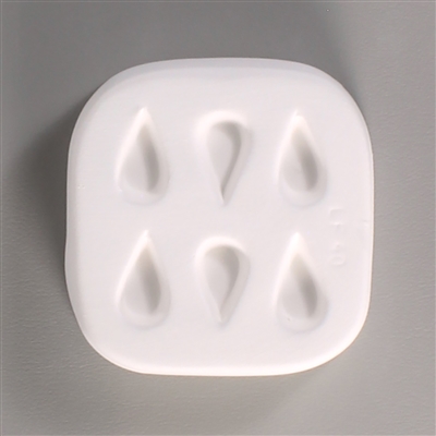 A square white ceramic mold for fusing hot glass on a grey background. Six small separate but identical teardrops have been carved into it with equal space between them.