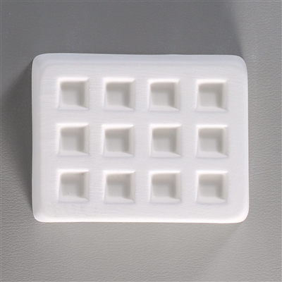 A rectangular white ceramic mold for fusing hot glass on a grey background. Twelve small separate but identical squares have been carved into it with equal space between them.