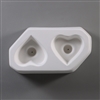 A white ceramic mold for fusing hot glass on a grey background. Two separate identical hearts have been carved into it facing opposite directions. Each heart has a deeper circle in the center with a small vent hole in the very center.