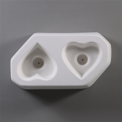 A white ceramic mold for fusing hot glass on a grey background. Two separate identical hearts have been carved into it facing opposite directions. Each heart has a deeper circle in the center with a small vent hole in the very center.