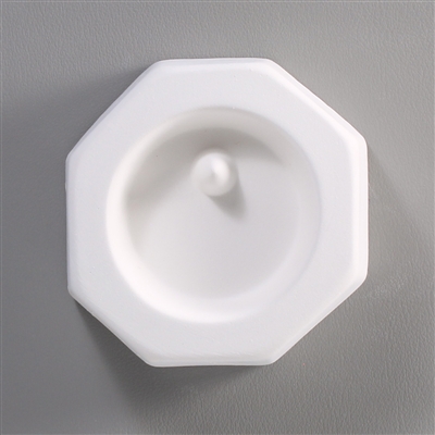 An octagonal white ceramic mold for fusing hot glass on a grey background. A circle has been carved into it. There is a post towards the top of the circle allowing for the resulting glass to be strung as jewelry.