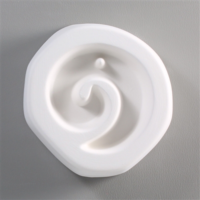 A roughly circular white ceramic mold for fusing hot glass on a grey background. A simple counterclockwise spiral has been carved into it. There is a post towards the top of the spiral allowing for the resulting glass to be strung as jewelry.