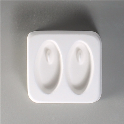 A square white ceramic mold for fusing hot glass on a grey background. Two identical long ovals have been carved into it. There is a post towards the top of each oval allowing for the resulting glass to be strung as jewelry.