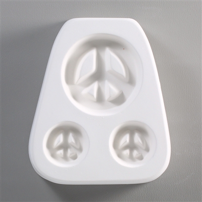 A white ceramic mold for fusing hot glass on a grey background. Three peace signs have been carved into it, one large above and two smaller below. Their empty spaces are left raised and uncarved allowing for the resulting glass to be strung as jewelry.