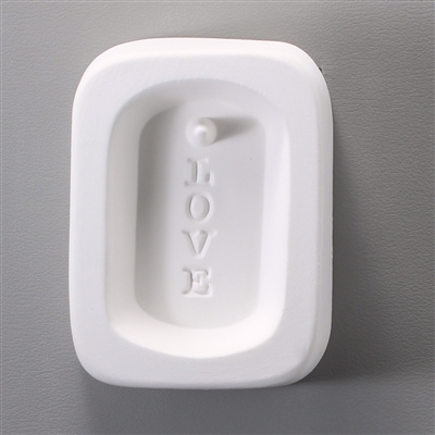A rectangular white ceramic mold for fusing hot glass on a grey background. An oval with the word LOVE vertically in block letters on it has been carved into it. There is a post at the top of the oval allowing for the final piece to be strung as jewelry.