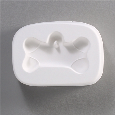 A rectangular white ceramic mold for fusing hot glass on a grey background. A dog bone shape has been carved into it. There is a post above the middle of the dog bone allowing for the resulting glass piece to be strung or otherwise hung.