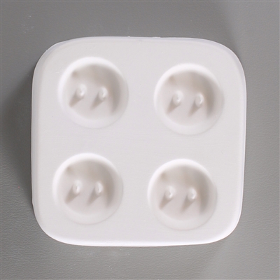 A square white ceramic mold for fusing hot glass on a grey background. Four identical small circular buttons, each with two posts to create holes for threading, have been carved into it with equal space between them.