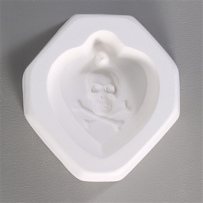 An octagonal white ceramic mold for fusing hot glass on a grey background. An inverted tear-shaped cameo style design of a skull and crossbones has been carved into it. There is a post above the skull allowing for the final piece to be strung.