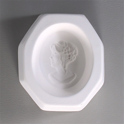 An octagonal white ceramic mold for fusing hot glass on a grey background. An oval-shaped cameo style design of a profile of a womanâ€™s face looking left has been carved into it. The woman has two small flowers in her hair by her ear.