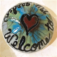 A dome of fused glass. There is a red heart outlined in black on the top, and light blue and green radiate outwards from it. There are traces of reactivity where blue and green meet. Welcome to My Garden has been written around the domeâ€™s edge in black.