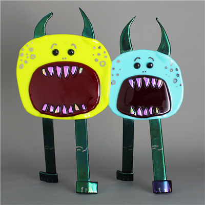 Two standing creatures made of fused glass. They each have a single round piece as their head/body, two curved horns, and two long legs. They each have large round mouths with small iridescent teeth and two small black eyes above it.