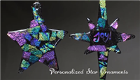 Two fused glass star ornaments hang in front of a dark grey background. The right has five points and the left eight. Both are black glass with squares of iridescent glass throughout reflecting cool toned colors. The right has joy written in the center.