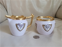 An antique Theodore Haviland Limoges porcelain creamer and open sugar bowl feature a soft pink background with gold and black medallions containing lotus floral designs alongside heavy gold rims and handles