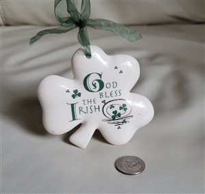 Preowned 1994 Russ Berrie porcelain shamrock hanging ornament with green ribbon that reads God Bless the Irish next to a quarter for size scale